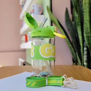 Clear water bottle with green lid and strap on a wooden surface, with a plant in the background.